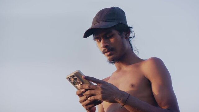 Daylight outdoor shot of a young shirtless Indian Asian male teenager wearing a cap using a mobile phone or smartphone to send a text message or use social media, against the blue sky in the evening. 