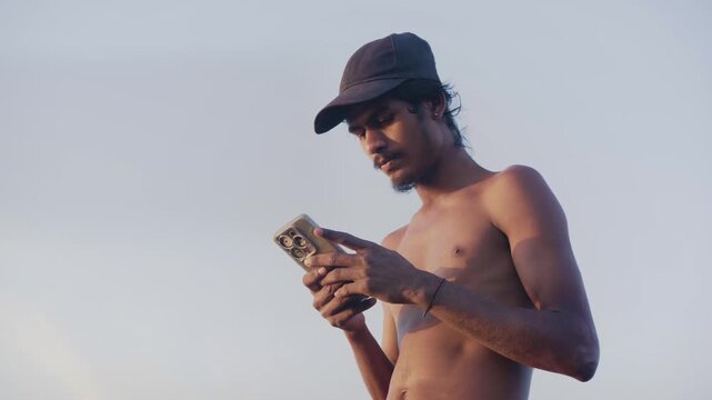 Daylight outdoor shot of a young shirtless Indian Asian male teenager wearing a cap using a mobile phone or smartphone to send a text message or use social media, against the blue sky in the evening. 