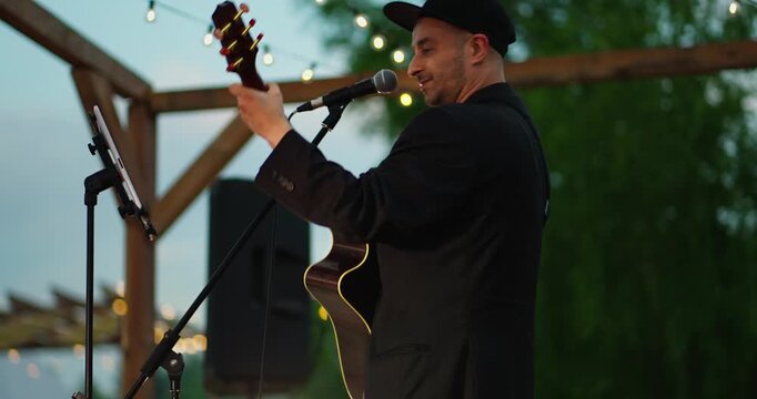 Male guitarist singing on wooden outdoor stage at dusk under string lights
