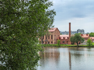 Obraz premium Old red brick factory building with chimney by a lake framed by willow tree