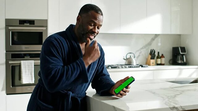 Man In Blue Morning Robe Using Smartphone With A Green Screen In A Bright Modern Kitchen With Marble Countertops