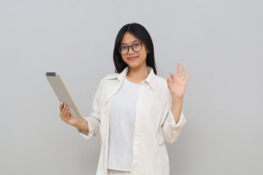 A young Asian woman holding a computer digital tablet while making an "okay" sign with her fingers, smiling looking at the camera, standing isolated on a white background.