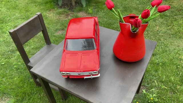 Retro Charm on Display: A vintage red toy car sits beside a vibrant red pitcher filled with blooming tulips, creating a whimsical still life on a wooden table in a garden setting.
