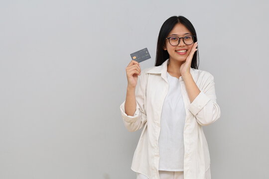 Photo of an impressed young Asian woman, hand on cheek, holding credit card, smiling looking at camera, isolated on white background.