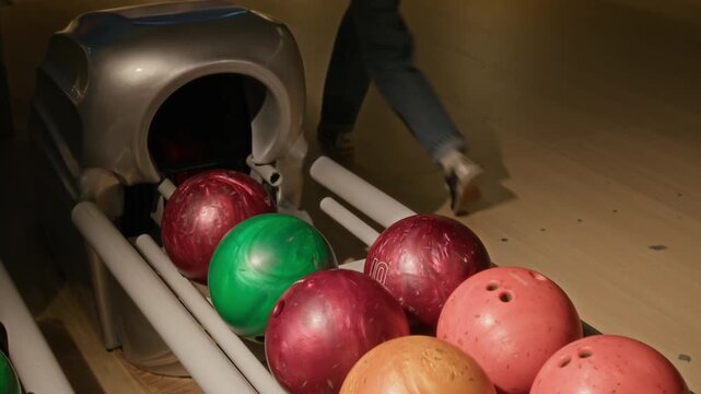 Young woman choosing and picking up orange bowling ball from return machine and walking away while playing with friends. Marketing indoor sports, weekend leisure, or entertainment venues