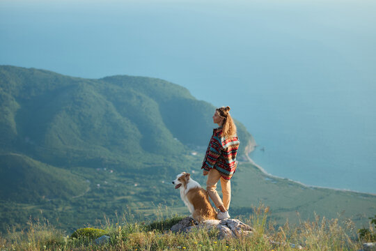 A woman stands beside her Border Collie overlooking mountain scenery and distant sea. Scenic surroundings create peaceful outdoor lifestyle atmosphere.