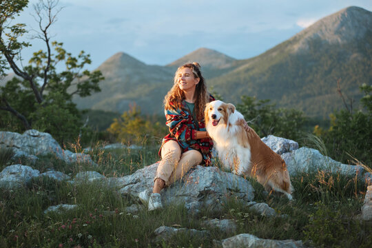 A woman sits beside her Border Collie overlooking mountain scenery and open sky. Peaceful surroundings create a calm travel atmosphere.