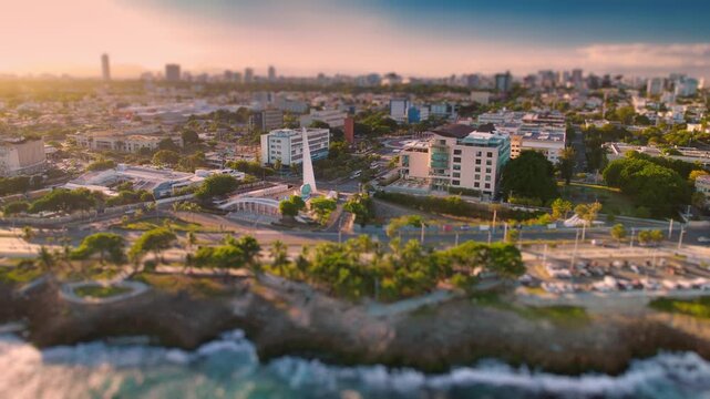 High-angle aerial drone shot of the Centro de los H&eacute;roes area and the coastal Malec&oacute;n in Santo Domingo, Dominican Republic, featuring a tilt-shift "miniature" effect during golden hour, transforming t