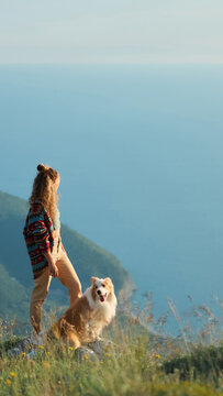 A woman walks with her Border Collie along a scenic mountain viewpoint above the coast. Outdoor scenery creates travel and adventure inspiration.