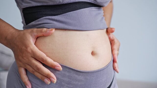 close-up of a woman in grey athletic wear pinching her belly fat, representing body weight struggles, fitness goals, healthy lifestyle changes, body positivity, and obesity concern