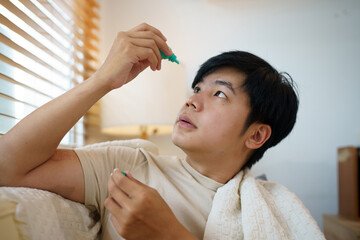 Naklejka premium Young Asian Man Applying Eye Drops at Home for Dry Eye Relief or Allergy Treatment
