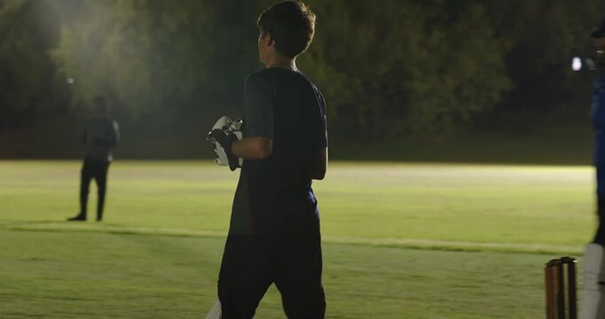 After helmet tossing, male batsman in blue-black jersey swinging bat on pitch, lights