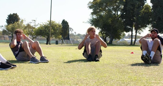 Performing sit-ups male teammates lying on grass under trees in park, training cones