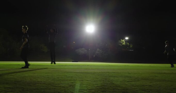 Bowler releasing ball for wicket, diverse male cricketers celebrating stumps, floodlit outfield