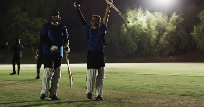 Two male batters celebrating win under floodlights raising bats, shaking umpire hand, walking