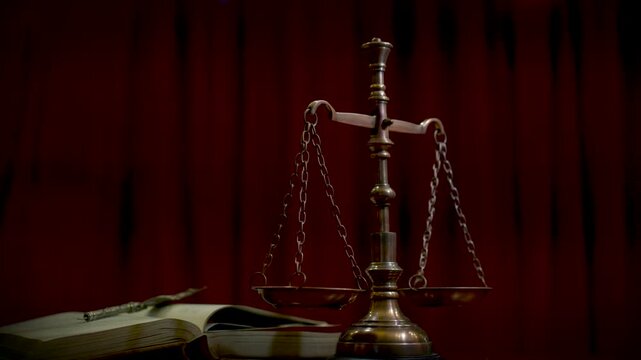 A brass scale of justice and aged law books arranged on a wooden desk with a dark red curtain background in warm dramatic lighting.