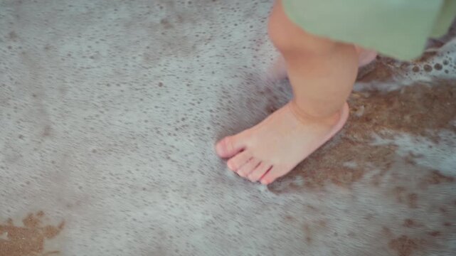 Close-up of an Asian toddler's feet standing on a sandy beach while white sea foam washes over them at sunset. Childhood discovery and sensory play during a family vacation in warm.