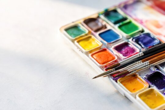 Watercolor paint palette with vibrant colorful pans and professional brushes resting on white textured surface in bright studio light, creative art concept with copy space