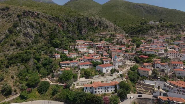 Aerial view of the traditional hillside village of Dhermi with white stone houses and red roofs nestled against lush green mountains in Dhermi, Vlore County, Albania.