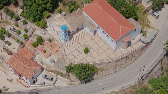 Aerial view of a church with a red roof and blue-domed bell tower featuring a paved courtyard and surrounding greenery in Dhermi, Vlore County, Albania.