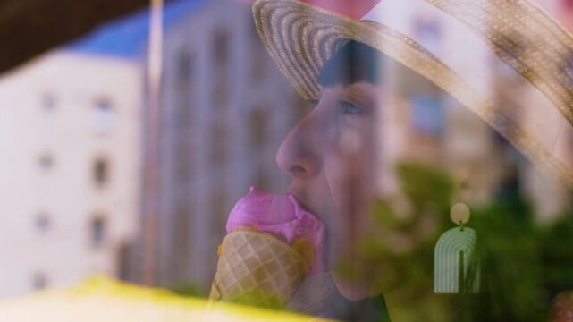 A young woman in a wicker straw hat and a pink patterned blouse smiles while holding an ice cream cone with two scoops of bright pink and orange ice cream inside. Summer mood.