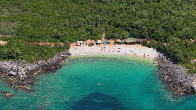 Aerial view of Aquarium Beach featuring turquoise waters, rocky cliffs, and lush green hills with campers on the sand in Jala, Vlore County, Albania.