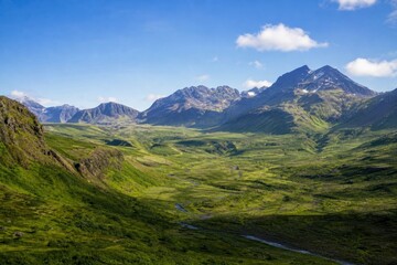Fototapeta premium Mountain valley with winding river flowing through green meadows under blue sky, majestic wilderness landscape, peaceful outdoor adventure background.