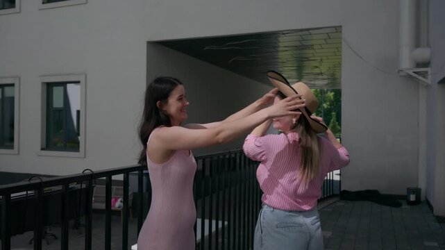 Two women share joyful, playful moments on a sunny balcony. One laughs as she playfully adjusts the other's hat. Their smiles and gestures convey lighthearted friendship. Perfect for scenes of casual