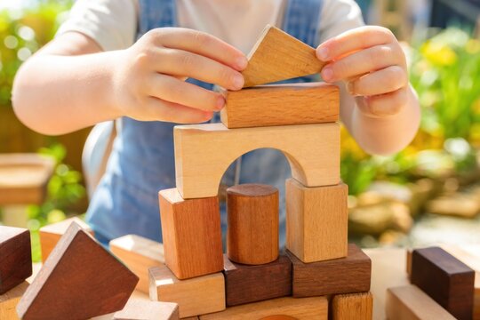 Child hands building wooden block tower in sunny outdoor garden, creative educational play, close-up shot with soft natural light