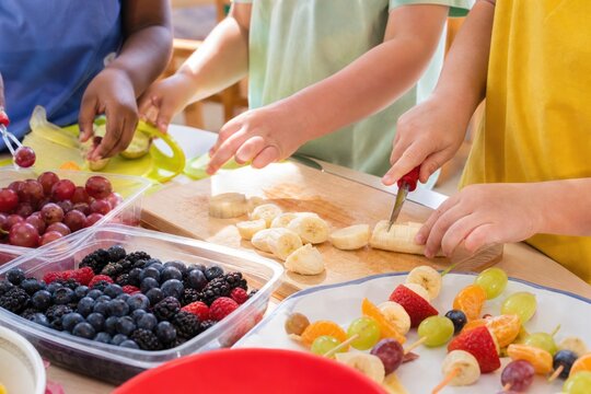 Diverse children hands slicing fresh organic banana on wooden board and preparing healthy fruit skewers in bright classroom cooking class, close-up view