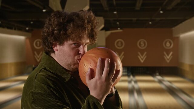 Portrait of mischievous red-haired Caucasian young gen Z man holding orange ball, kissing for luck and smiling at camera in bowling alley. Perfect for sports marketing or leisure venue promos