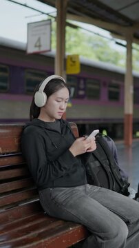 Traveler sitting on a bench using smartphone with headphones at a train station, ideal for travel lifestyle, waiting, mobile app, music and railway journey concepts