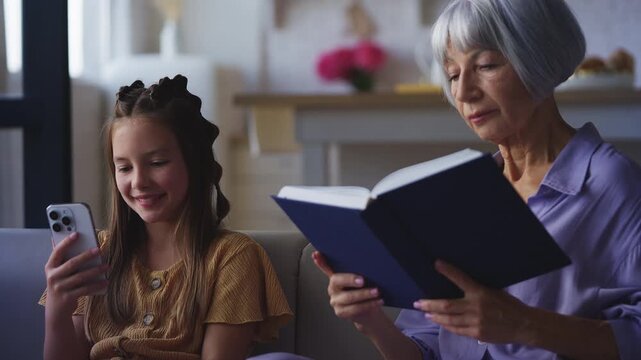Focused granny reading a book while her granddaughter texts on a phone, hobbies