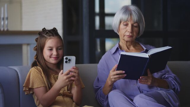 Happy girl using a phone while her grandmother reads a book, relaxing at home