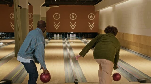 Black young man and Caucasian friend clinking bowling balls and throwing down lanes in alley together during team tournament. Clip is ideal for marketing campaigns on friendship and active recreation