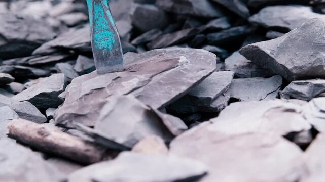 Slow-motion macro shot. A chisel is placed on granite. Hammer blows strike the tool, causing tiny stone particles and fine dust to erupt from the point of impact with each strike.