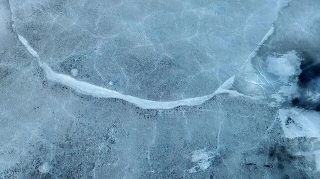 Graphic, top-down aerial shot of a deep fissure running through a sheet of thick blue ice during the spring thaw cycle.