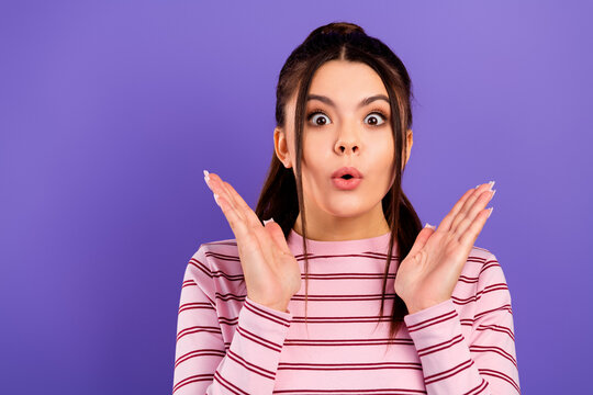 Surprised young woman with hands raised and expressive face on purple background, casual striped pink shirt portrait showing shock, amazement and playful emotion