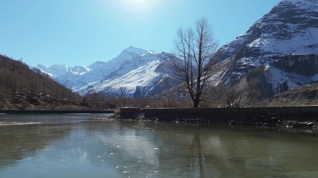 Sissu Lake in Lahaul Valley reflects snow-covered Himalayan mountains under clear blue sky. Static wide angle shot captures partially frozen glacial lake with bare winter trees along shoreline.