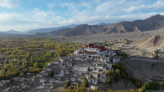 Thikse Monastery perched on a hilltop in the Indus Valley, surrounded by traditional Tibetan Buddhist architecture and green agricultural fields. Aerial perspective captures the monastery complex.