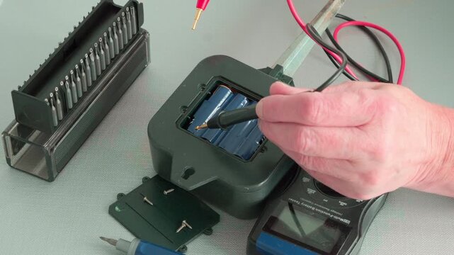 Hands test battery in device with multimeter. Tools and wires surround the setup. Scene shows practical electronics maintenance. Footage captures focused, hands-on technical work

