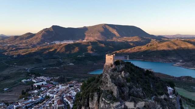 Aerial view of Zahara de la Sierra Castle on a rocky peak overlooking the white village and turquoise reservoir under golden hour light in Zahara de la Sierra, Andalucia, Spain.