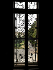 Obraz premium Dark interior view through an ornate window with decorative wrought iron grille overlooking a quiet garden outside.