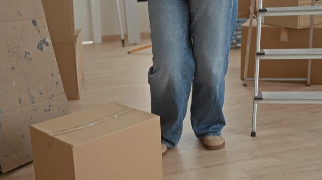 Young blonde woman in oversized blue sweater dancing while unpacking moving boxes beside a stepladder, arms raised in a sunlit building; joy celebration.