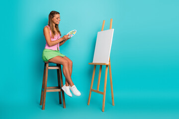 Fototapeta premium Young woman painting on a canvas in a bright blue studio background with a smile and summer chic vibe