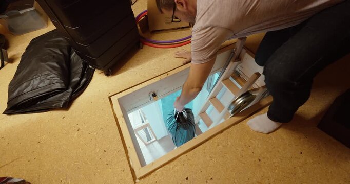 Person passing a bag down through an attic hatch using the loft access opening and ladder.