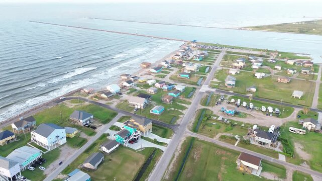 Shoreline stilt rows frame homes along Seashell Dr inland from Surfside Beach waterfront where jetty wave line track coast. Varied siding tones, raised decks define block in Freeport, Texas