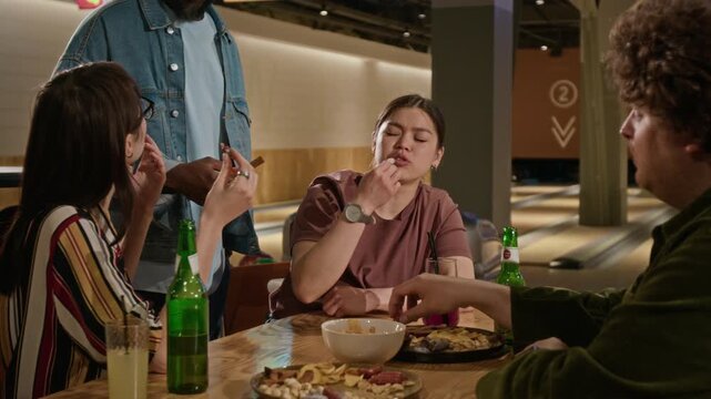 Diverse young zoomer friends sitting at table in bowling alley, eating snacks, drinking beer, and talking. Perfect for commercial use in hospitality, entertainment, and social lifestyle campaigns