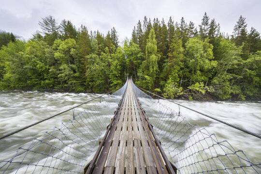 View of a wooden suspension bridge crossing a rushing river surrounded by lush green forest in Junkerdal, Nordland, Norway.
