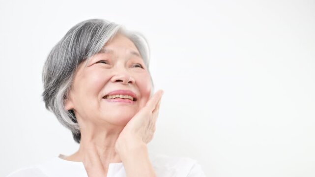Beauty concept video featuring a close-up of an elderly woman (senior) with gray hair against a white background, looking up with a smile and touching her cheek with her hand